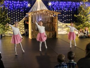 Trzy uczennice tańczą na tle szopki betlejemskiej. Dziewczynki mają wyciągnięte lekko do tyłu lewe nogi a prawe dłonie podnoszą ku górze.