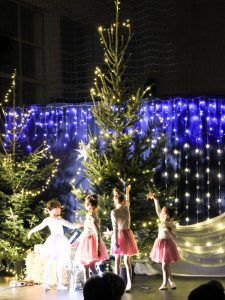 Cztery dziewczynki w strojach balerin stoją przy choince wyciągając w jej stronę ręce z gwiazdkami. W tle widoczna jest dekoracja z lampek umieszczona pionowo w rzędach na tle niebieskiego materiału.