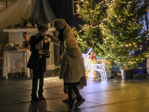 Zdjęcie przedstawia scenę ze szkolnych jasełek. Kobieta wraz z małą dziewczynką stojącą u jej boku odbiera z rąk chłopca stojącego na przeciwko małą latarenkę.