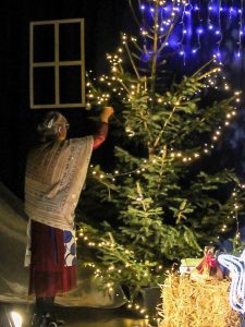 Zdjęcie przedstawia scenę ze szkolnych jasełek. Kobieta w beżowej chuście zarzuconej przez plecy wiesza na choince ozdobę.