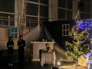 Zdjęcie przedstawia scenę ze szkolnych jasełek. Kobieta siedzi przy stole zakrytym białym obrusem. Za nią widoczny jest biały kominek. Obok stoją dwie uczennice trzymające podkładki z tekstem przed sobą. Po prawej stronie widoczna jest choinka.