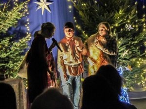 Zdjęcie przedstawia scenę ze szkolnych jasełek. Kobieta lekko pochyloną, stojące bokiem mówi do trzymanego przed sobą mikrofonu. Obok niej stoi dwóch uczniów patrzących w jej kierunku. Jeden z nich ma zawieszone przez ramię futro.