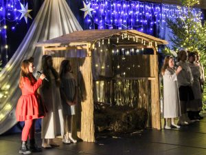 Wokół ustawionej na scenie szopki stoją uczennice. Dwie z nich trzymają mikrofony. Wszystkie dziewczynki ubrane są na biało poza jedną, która jest w czerwonym stroju.