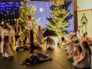 Zdjęcie przedstawia scenę ze szkolnych jasełek. Dziewczynki przebrane za aniołki siedzą w pół okręgu. Po środku stoi bokiem nauczycielka z mikrofonem w dłoni, a przed nią leży na ziemi uczennica w jasno fioletowej sukience.