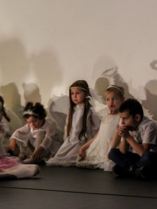 Czworo dzieci - trzy dziewczynki przebrane za aniołki i chłopiec siedzą na podłodze i patrzą przed siebie na przedstawienie.