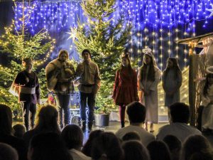 Sześcioro uczniów stoi na tle dwóch choinek oraz światełek zawieszonych na niebieskim materiale za nimi. Dzieci są ubrane w stroje w których występowały. Jeden z chłopców trzyma przed sobą mikrofon.
