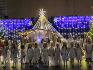 Dziewczynki ubrane w stroje aniołkoów stoją w rzędzie. Część z nich trzyma się za ręce. W tle widać dekorację przedstawienia - szopkę za którą zawieszony jest biały materiał zwieńczony na górze świecącą gwiazdą a za nią na niebieskim tle zawieszone w pionowych rzędach lampki w kształcie gwiazdek.