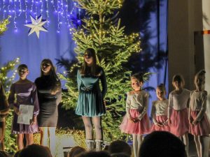 Na tle choinki w lekkim półcieniu stoją w kolorowych jednolitych sukienkach trzy uczennice klasy VII oraz cztery ich młodsze koleżanki w białych bluzeczkach i różowych tiulowych spódniczkach.