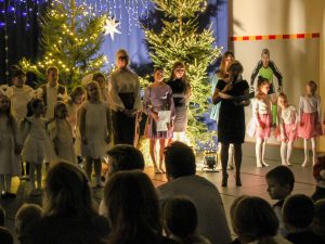 Pani dyrektor przemawia podczas jasełek. Kobieta trzyma w jednej dłoni mikrofon aa w drugiej rozłożoną teczkę z tekstem przemówienia. W tle widać stojące dzieci biorące udział w jasełkach oraz nauczycielkę, która ich przygotowywała.