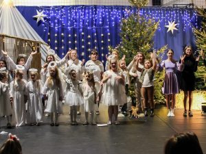 Uczniowie biorący udział w przedstawieniu podczas śpiewania kolędy. Dzieci ubrane na biało, oraz dwie dziewczynki w jednolitych sukienkach podnoszą w górę ręce.