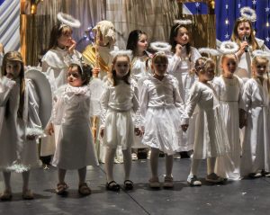 Dziewczynki w strojach aniołków stoją w rzędzie na scenie. Za nimi widoczny jest fragment szopki oraz czworo uczniów w niej stojących.