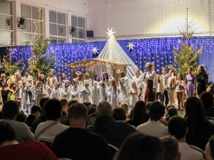 Uczniowie biorący udział w jasełkach stoją razem na scenie. Obok nich stoi nauczycielka trzymająca mikrofon w dłoni. W tle widać dekoracje - szopkę, choinki i lamki a przed nimi fragment widowni.