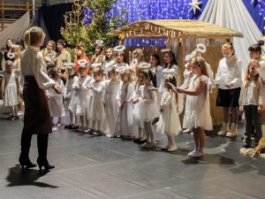 Przed ubranymi na biało dziewczynkami z aureolami na głowach stoi nauczycielka dyrygująca chórem. W tle widać dekoracje w postaci szopki betlejemskiej, choinek i zawieszonych światełek.