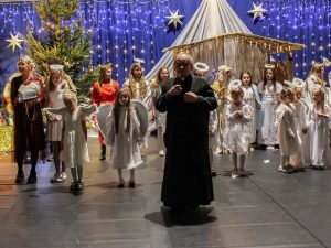 Na tle dzieci występujących w jasełkach stoi proboszcz trzymający w dłoni mikrofon.
