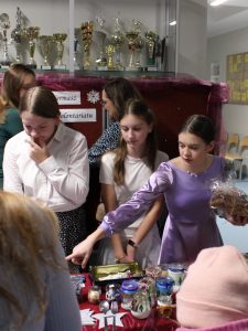 Trzy dziewczynki stoją za stołem z rozstawionymi przedmiotami na sprzedaż w świątecznym kiermaszu. Jedna z dziewczynek trzyma w dłoni foliową paczkę, a prawą ręką wskazuje na jakiś przedmiot. Jej koleżanki patrzą w stronę stołu.