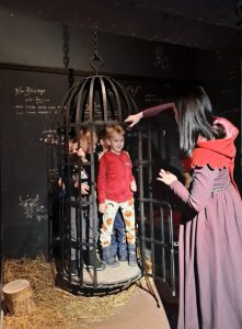 Oddział przedszkolny w "domu Legend". Uczniowie wychodzą z zawieszonej nad ziemią klatki. Kobieta ubrana w długą bordową suknię trzyma otwarte drzwi klatki i patrzy w stronę dzieci.