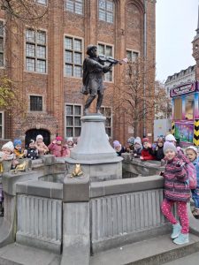 Uczniowie oddziału przedszkolnego stoją wokół fontanny z ustawionym w centralnym miejscu pomnikiem skrzypka. Dookoła fontanny na cokołach ustawione zostały złote żabki.