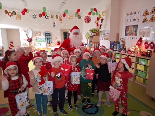 Uczniowie oddziału przedszkolnego pozują do wspólnego zdjęcia wraz z mężczyzną przebranym za Świętego Mikołaja. Dzieci mają na głowach czerwone czapeczki a w dłoniach trzymają papierowe torebki z prezentami.