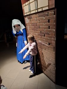 Oddział przedszkolny w "domu Legend". Uczennica stoi przy pochyłej ścianie z cegieł. Dziewczynka ma wyciągnięte przed sobą ręce. Obok stoi kobieta w długiej niebieskiej spódnicy i białej chuście na głowie. Pani wskazuje palcem skierowanym w dół na ścianę. Dziewczynka patrzy w jej stronę.