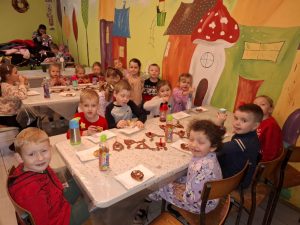 Uczniowie oddziału przedszkolnego siedzą przy długich stołach. Przed nimi leżą papierowe talerzyki, na których leża pączki. Obok talerzyków ustawione są butelki z napojami. Dzieci z pierwszego stolika patrzą w stronę aparatu i się usmiechają.