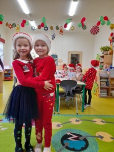 Dwie uczennice oddziału przedszkolnego pozują do wspólnego zdjęcia obejmując się na wysokości pasu. Dziewczynki uśmiechają się. Za nimi widoczni są pozostali uczniowie siedzący przy wspólnym stole. Nad sufitem wisi kolorowa girlanda z motywem świątecznym.