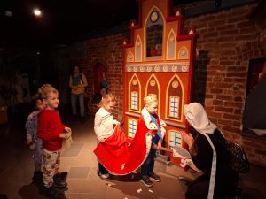 Oddział przedszkolny w "domu Legend". Uczniowie biorą udział w przedstawieniu. Jeden z chłopców ma na głowie koroną, a na ramionach długi płaszcz króla, który podtrzymuje jego koleżanka stojąca za nim. Przed chłopcem klęczy kobieta ze złożonymi przed sobą dłoniami. Obok stoi dwóje uczniów przyglądających się scenie. W tle widoczna jest ściana z cegieł a na jej tle ustawiona dekoracja przedstawiająca ścianę zamku.