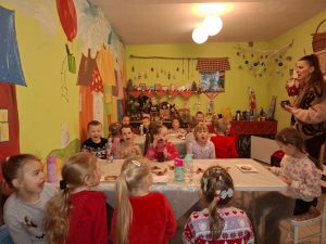 Uczniowie oddziału przedszkolnego siedzą w sali przy długich stołach. Ściany pomieszczenia są jasnozielone i ozdobione rysunkami i ozdobami umieszczonymi na półeczkach. Przed dziećmi stoi nauczycielka mówiąca w ich kierunku.