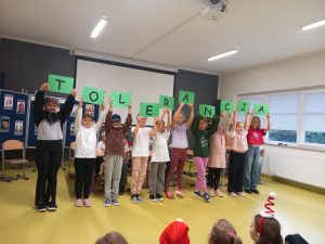 Uczniowie biorący udział w przedstawieniu stoją w jednym rzędzie. Każde dziecko trzyma w wyciągniętych nad głową dłoniach zieloną kartkę z nadrukowaną literą. Litery tworzą napis "TOLERANCJA".