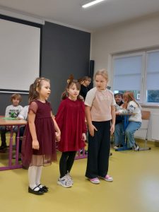 Trzy dziewczynki stoją w rzędzie podczas przedstawienia. Dwie z nich trzymają się za ręce a ich koleżanka patrzy w ich stronę.