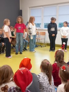 Zdjęcie pięciu uczennic podczas przedstawienia. Dziewczynki stoją w małych odstępach od siebie. Zarówno dwie znajdujące się po lewej jak i po prawej stronie patrzą się na siebie. Dziewczynka stojąca po środku samotnie patrzy w stronę swoich koleżanek.