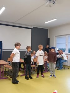 Trzech chłopców i dziewczynka stoją w rzędzie w małych odstępach od siebie. Chłopcy patrzą w stronę dziewczynki, która ma na głowie czapeczkę z daszkiem. Dziewczynka patrzy na chłopców.