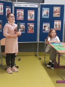 Uczennica grająca w przedstawieniu rolę nauczycielki stoi z podniesioną w górę głową. Trzyma w zaplecionych przed sobą dłoniach sztywną podkładkę. Obok za stolikiem , bokiem do niej siedzi inna uczennica. W tle za dziewczynkami widoczna jest niebieska tablica z przyczepionym u góry napisem "Zostań z nami tolerancjo..." a pod nim w odstępach przyczepione są kolorowe obrazki.