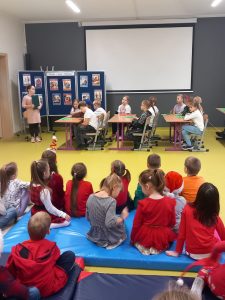 Zdjęcie przedstawia uczniów biorących udział w przedstawieniu. Dzieci siedzą w ławkach a przed nimi stoi ich koleżanka odgrywająca rolę nauczycielki. Na pierwszym planie widoczne są dzieci siedzące na materacach, które oglądają przedstawienie.