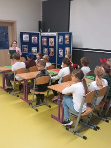 Uczniowie biorący udział w przedstawieniu siedzą w trzyosobowych ławkach ustawionych w trzech rzędach. Przed nimi stoi ich koleżanka odgrywająca rolę nauczycielki. Po jej prawej stronie ustawiona jest granatowa tablica z przyczepionymi kolorowymi obrazkami.