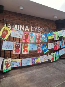 Zdjęcie wywieszonych w trzech rzędach obok siebie prac plastycznych przedstawiających misie. Za pracami widoczna jest ściana z cegieł, na którym umieszczony jest napis "GMINA ŁYSOMICE".