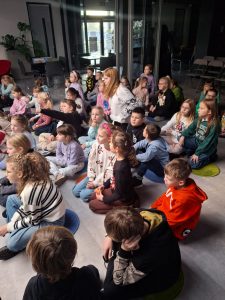 Uczniowie siedzą na podłodze w trakcie spotkania autorskiego. Dzieci patrzą przed siebie.