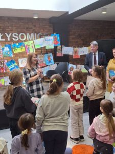 Uczniowie stoją na przeciwko autorki, która trzyma w dłoniach przed sobą mikrofon.