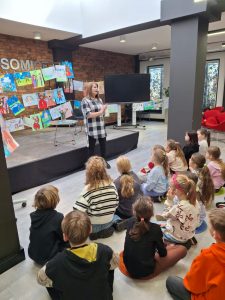 Uczniowie siedzą na podłodze sali spotkań Gminy Łysomice. Dzieci patrzą w stronę stojącej przed nimi autorki spotkań.