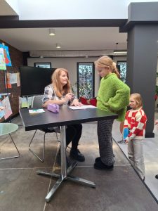 Uczennica stoi przy stole za którym siedzi autorka spotkania. Dziewczynka spogląda w dół na stół, a kobieta patrzy w jej kierunku.