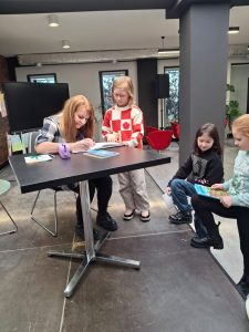 Uczennica stoi obok stołu za którym siedzi autorka spotkania. Kobieta pochyla się nad kartką z długopisem w dłoni, a dziewczynka patrzy w jej kierunku.