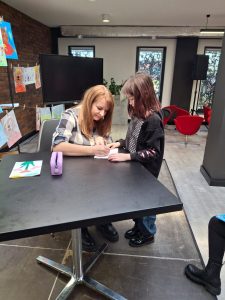 Uczennica stoi przy stole za którym siedzi autorka spotkania. Kobieta trzyma w dłoni długopis i pochyla się z nią nad kartką, która w dłoniach trzyma uczennica.