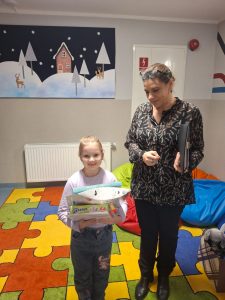 Uczennica stoi obok Pani bibliotekarki na dywanie. Dziewczyna trzyma w dłoniach przed sobą otrzymane w konkursie nagrody.