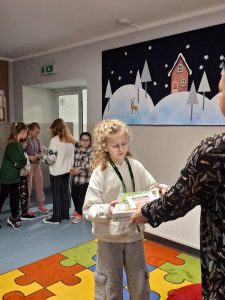 Uczennica odbiera nagrody w konkursie. Dziewczynka spogląda na nie. Na zdjęciu widoczne są dłonie nauczycielki je wręczającej. W tle widać stojące inne dzieci.