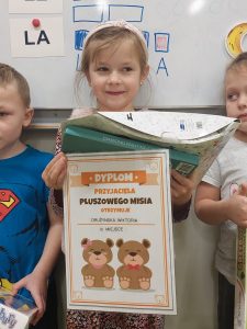 Uczennica stoi pod tablicą i szeroko uśmiecha się. W ręku trzyma dyplom oraz otrzymane w konkursie nagrody.