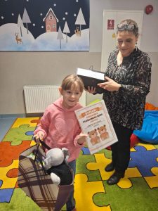 Uczennica odchodzi od Pani bibliotekarki, która wręczyła jej nagrody. Dziewczynka trzyma w jednej dłoni dyplom a w drugiej torbę z nagrodami, z której wystaje pluszowa zabawka.