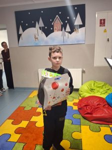 Uczeń stoi na kolorowym dywanie. Chłopiec trzyma przed sobą białą teczkę ozdobioną kolorowymi rysunkami.