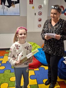 Uczennica stoi obok Pani bibliotekarki, która patrzy na nią z uśmiechem. Dziewczynka trzyma w dłoniach przed sobą nagrody.