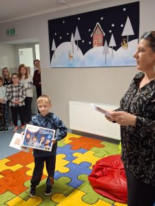 Uczeń stoi na kolorowym dywanie trzymając przed sobą duże pudełko - nagrodę otrzymaną w konkursie. po prawej stronie na skraju zdjęcia widoczna jest Pani bibliotekarka a w tle stojące inne dzieci.