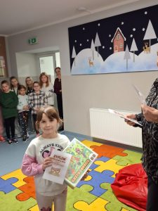 Uczennica pozuje do zdjęcia trzymając przed sobą otrzymaną w konkursie nagrodę oraz dyplom. Dziewczynka jest uśmiechnięta. W tle widać innych stojących uczniów.