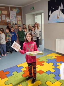 Uczennica pozuje do zdjęcia stojąc na kolorowym dywanie. Dziewczynka w jednej dłoni trzyma dyplom który podnosi w górę, a w drugie otrzymaną nagrodę. W tle widać stojące inne dzieci.